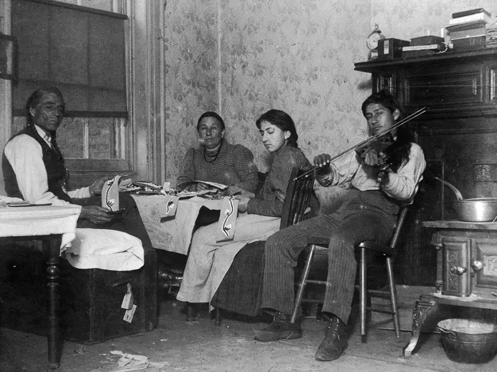 Mountain Eagle And His Family Make Native American Handicrafts While His Son Plays Violin In Their Tenement At 6 Beach Street, 1895.