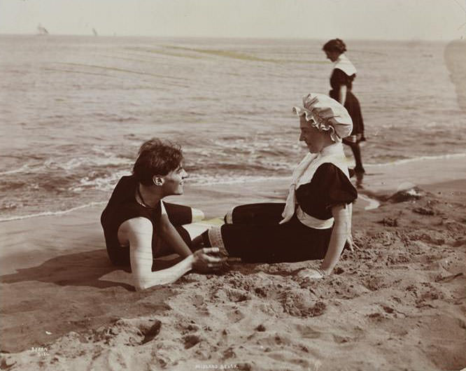 A Man And A Woman Lying On Midland Beach. Another Woman Stands At The Edge Of The Water.