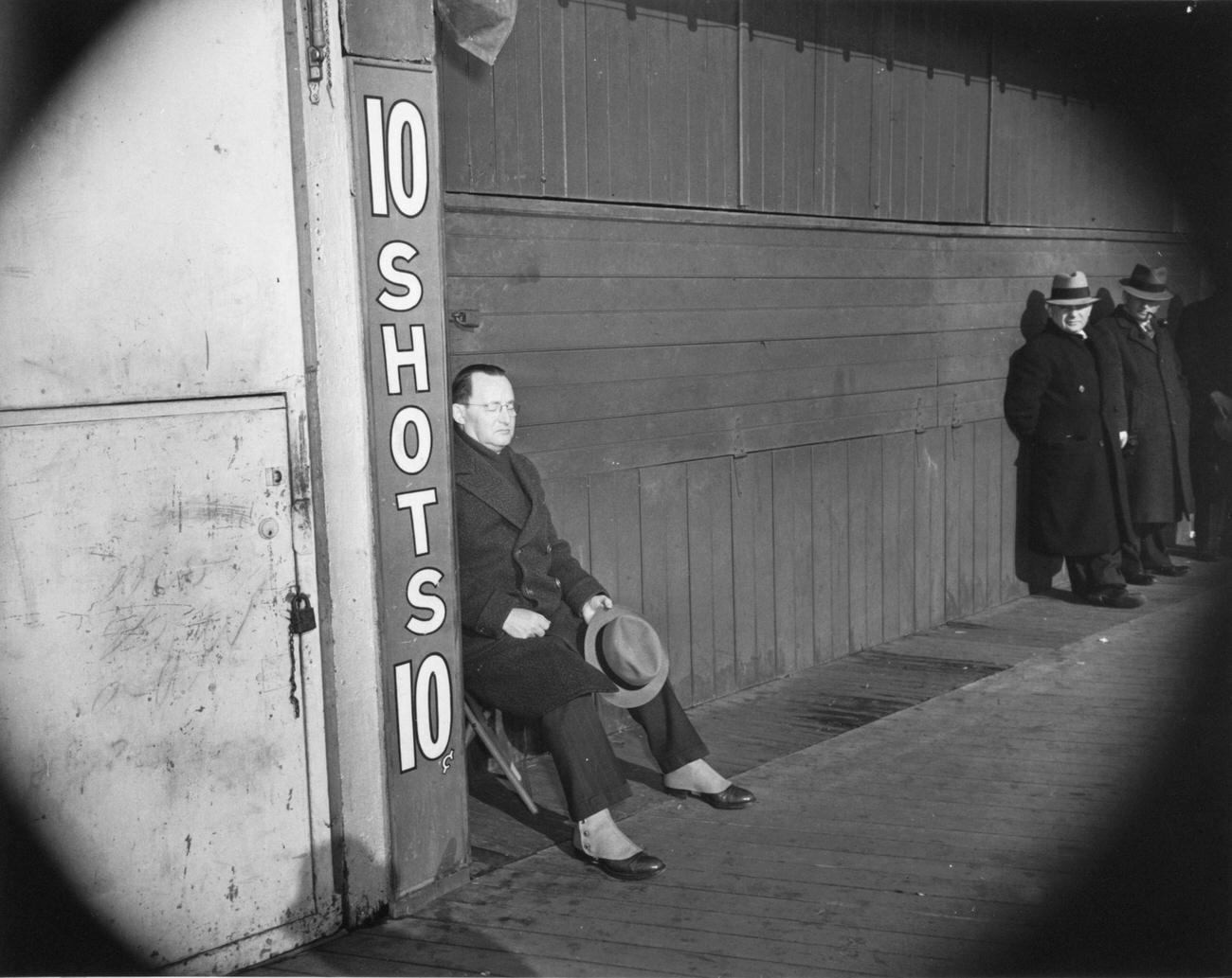 Man Sits In Folding Chair In Front Of Closed Shooting Gallery Gate, 1941