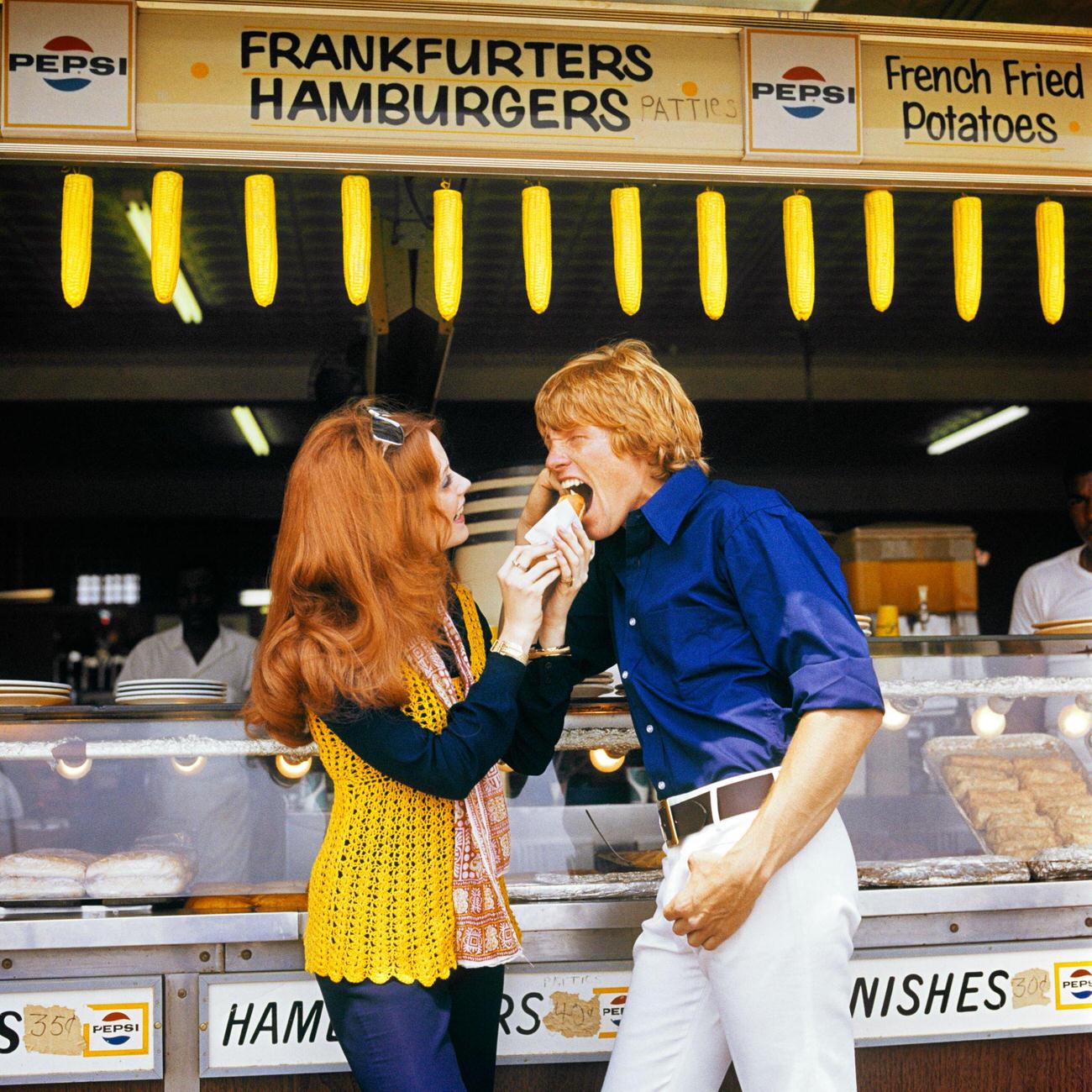Woman Feeding Man At A Coney Island Hot Dog Stand, 1970S