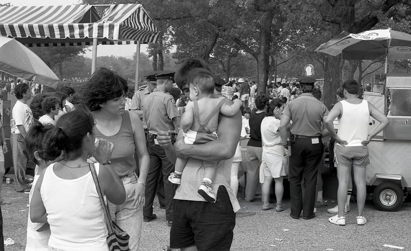 A Pair Of Women And A Young Boy Look At A Baby In The Arms Of A Man Near Vendor Stalls In Flushing Meadows Park, Corona, Queens, 1988.