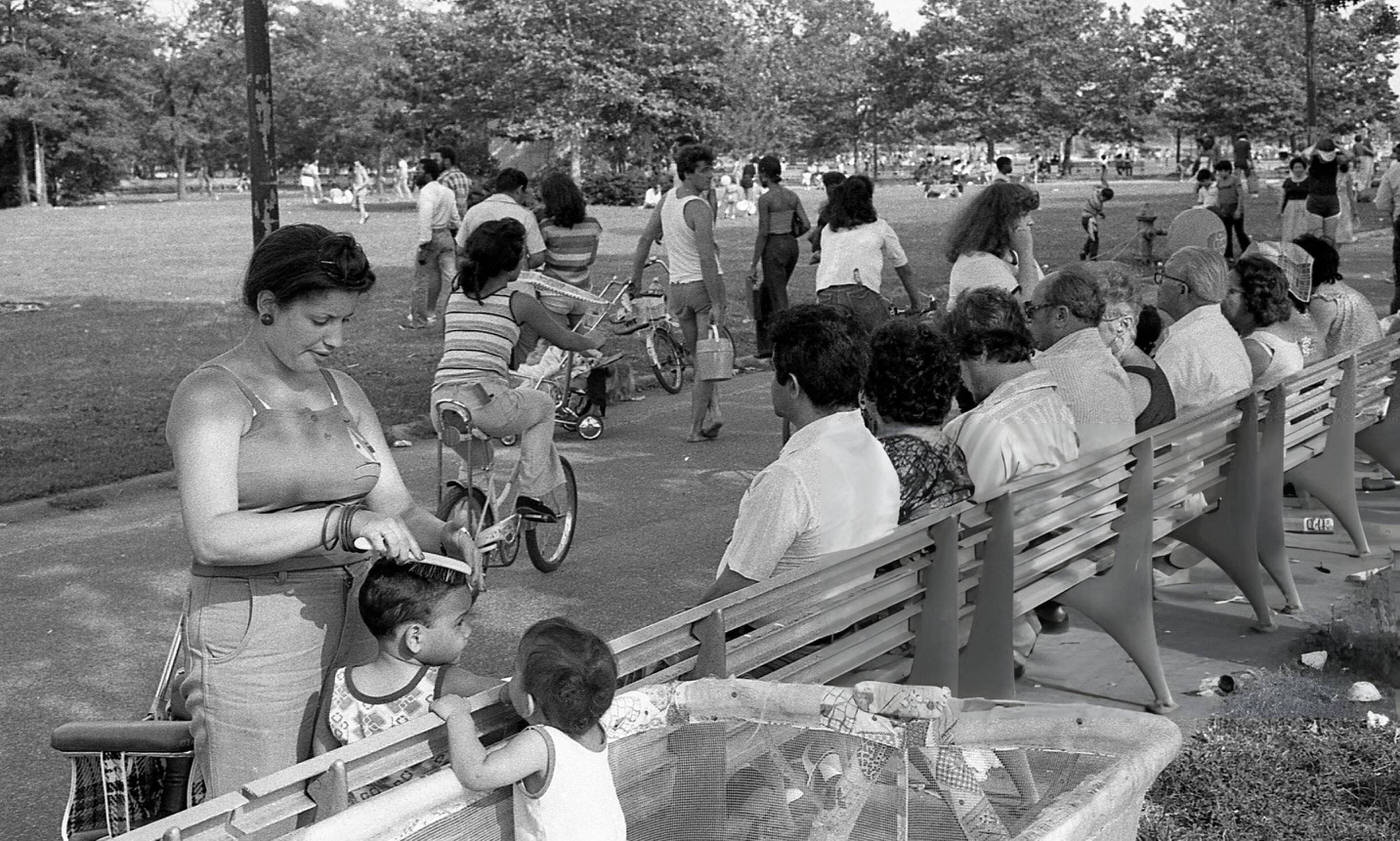 People sit on a park bench as a woman combs a child's hair in Flushing Meadows Park, Corona, Queens, 1980.