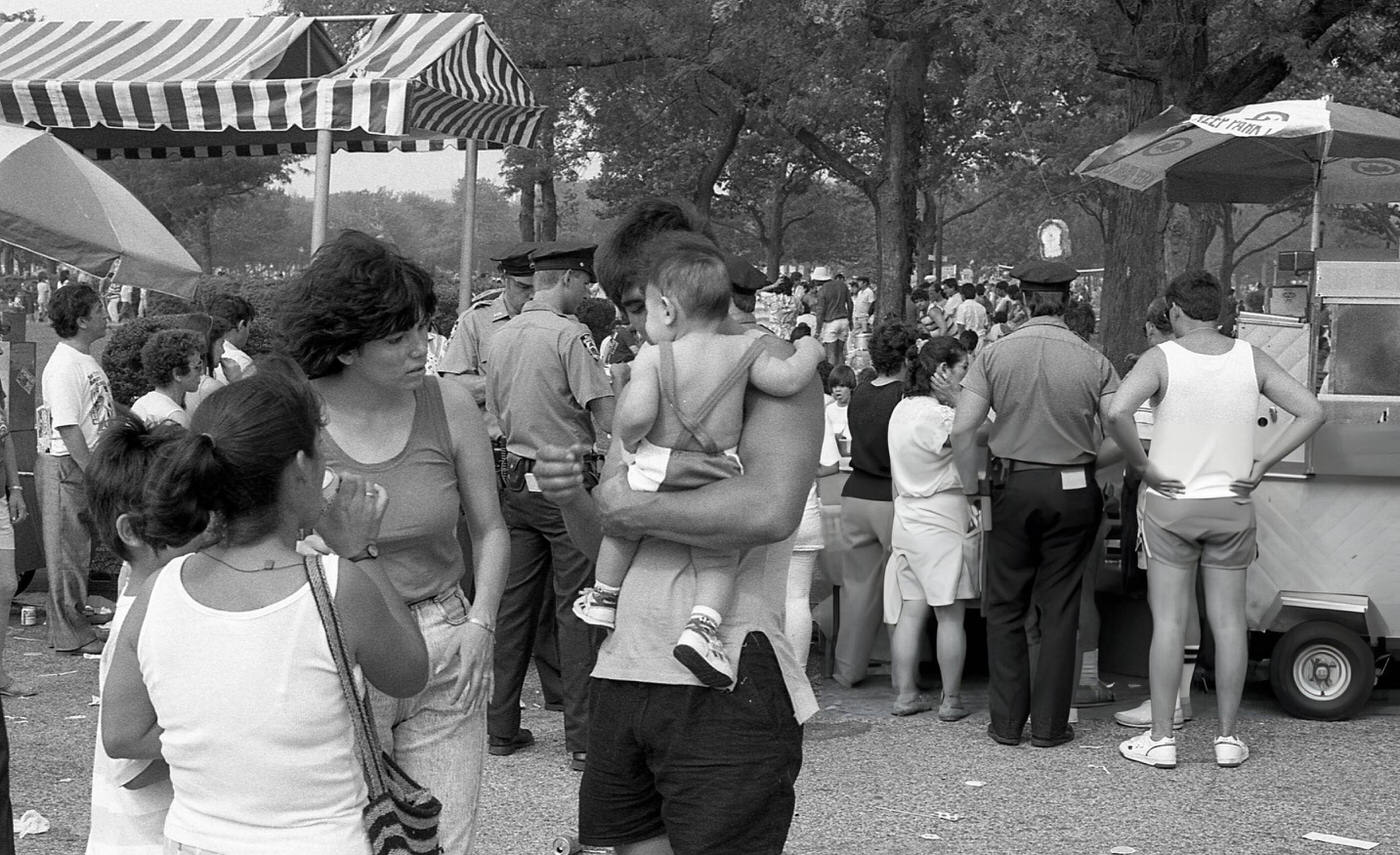 A pair of women and a young boy look at a baby in the arms of a man near vendor stalls in Flushing Meadows Park, Corona, Queens, 1988.