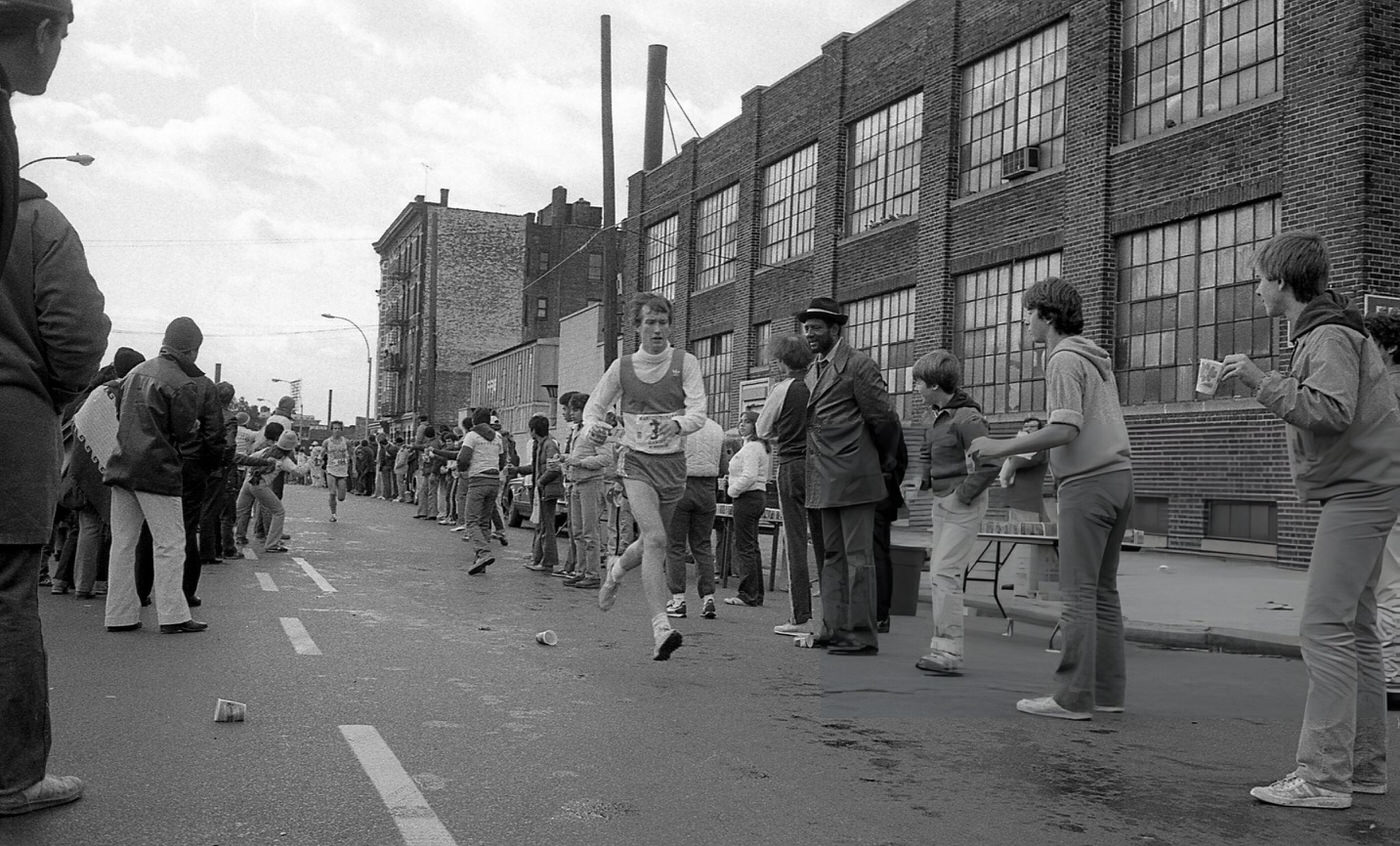 Spectators And Water Station Volunteers Line Crescent Street As A Runner Passes The 14-Mile Marker During The New York City Marathon, Queens, 1980.