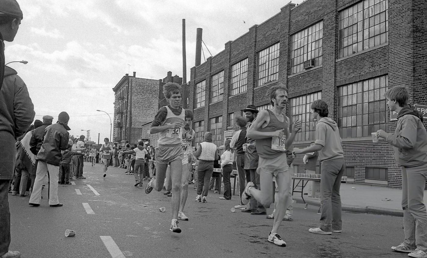 Spectators And Water Station Volunteers Line Crescent Street As Two Runners Pass The 14-Mile Marker During The New York City Marathon, Queens, 1980.