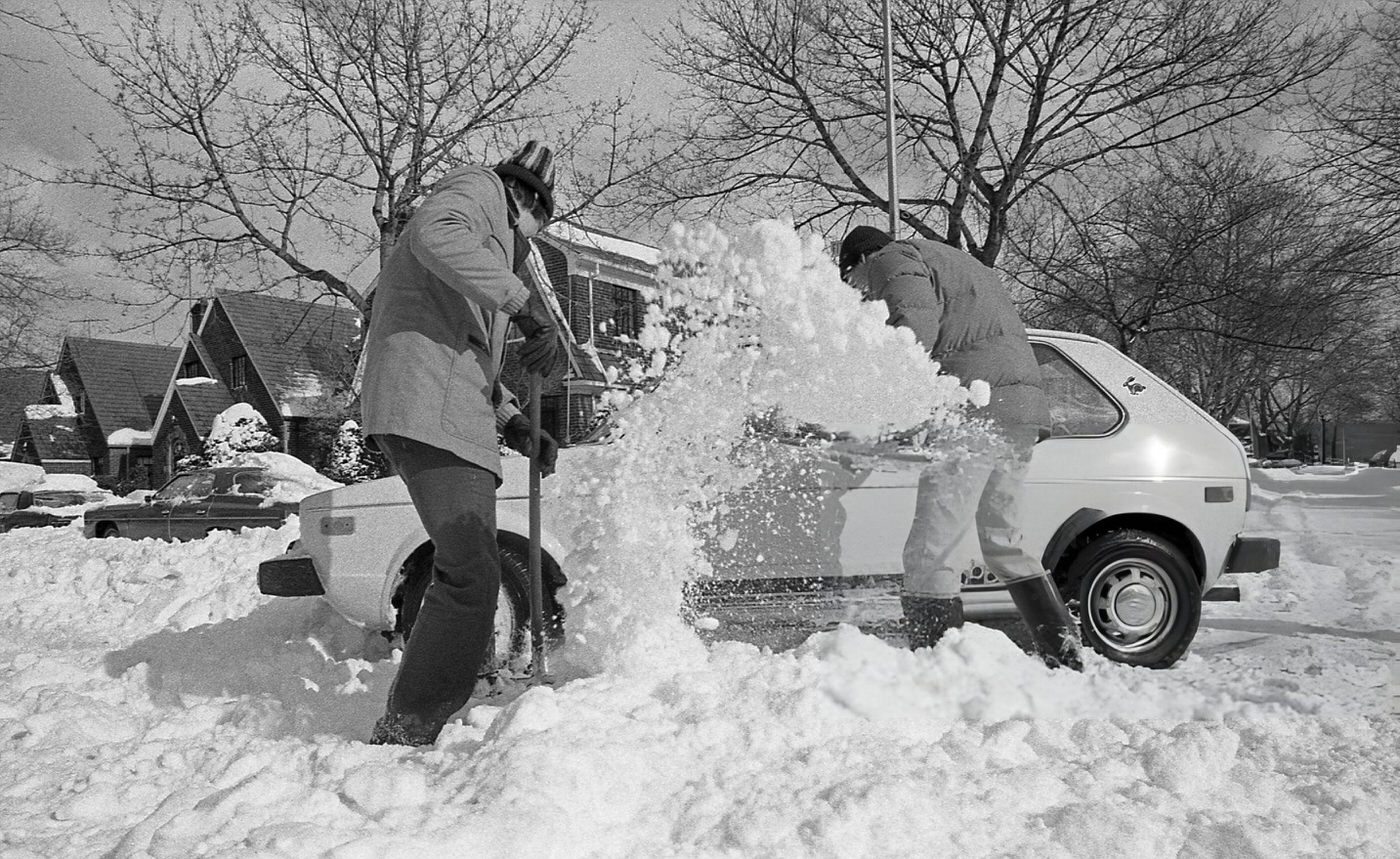 Two Men Digging Out A Volkswagen Rabbit Stranded In Deep Snow In A Residential Intersection In The Aftermath Of The Blizzard Of 1978.
