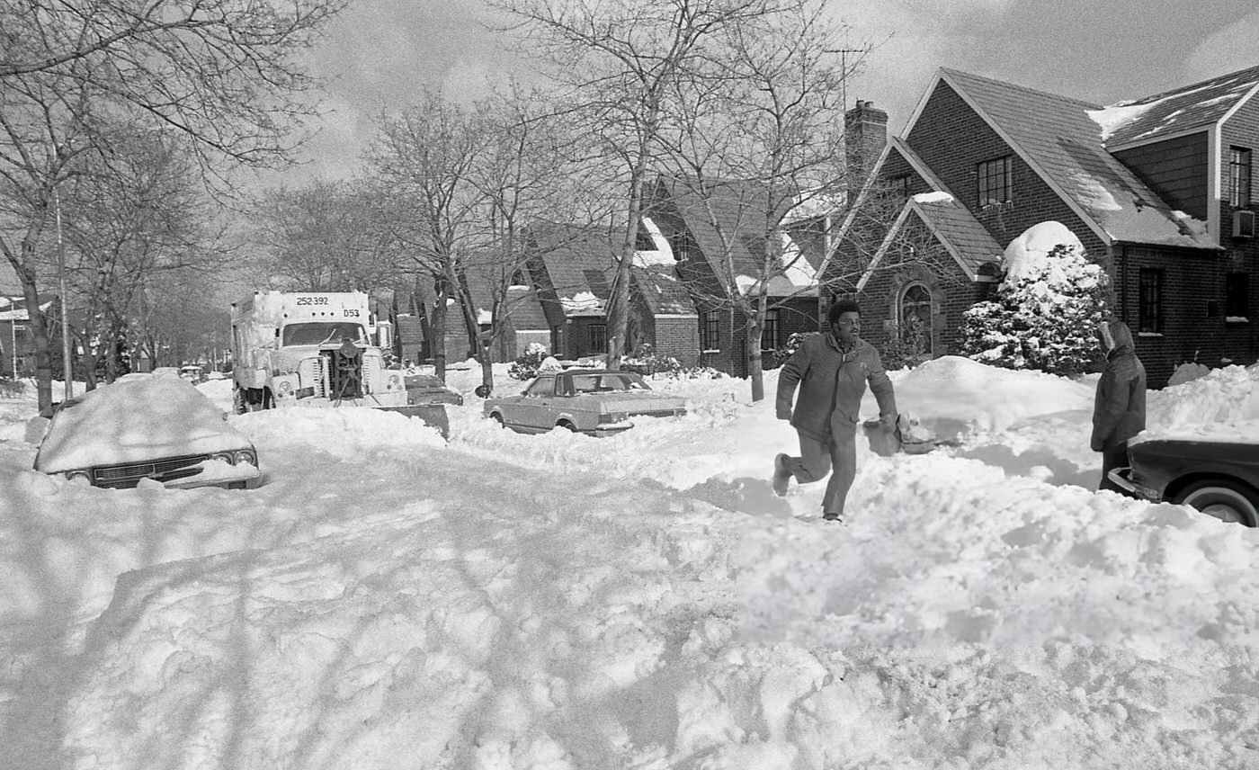A Man Runs Through Deep Snow On A Residential Street, As A Nyc Sanitation Snow Plow Pulls Up Behind Him In The Aftermath Of The Blizzard Of 1978.