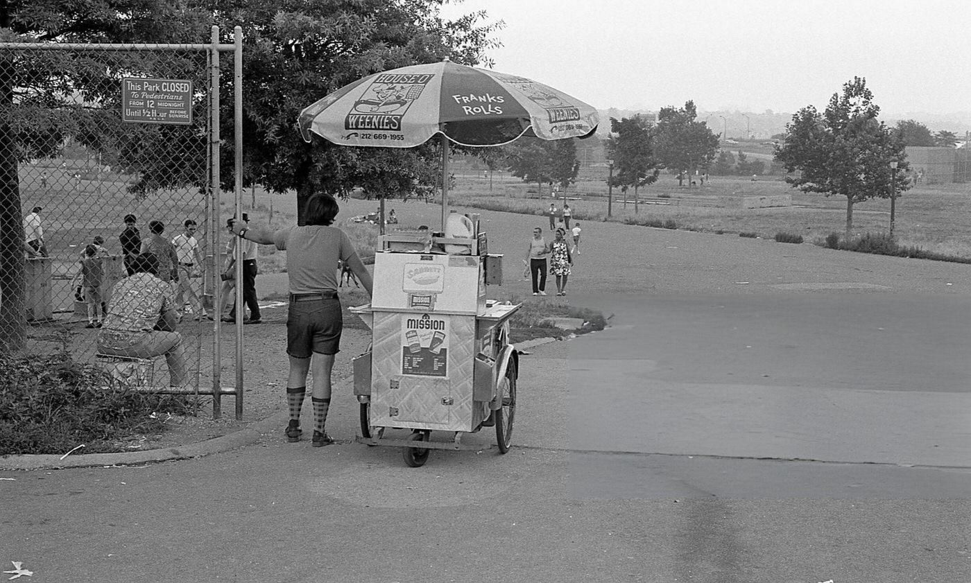 A Hot Dog Vendor Standing Beside His Cart In Flushing Meadows Park, Queens, 1975.