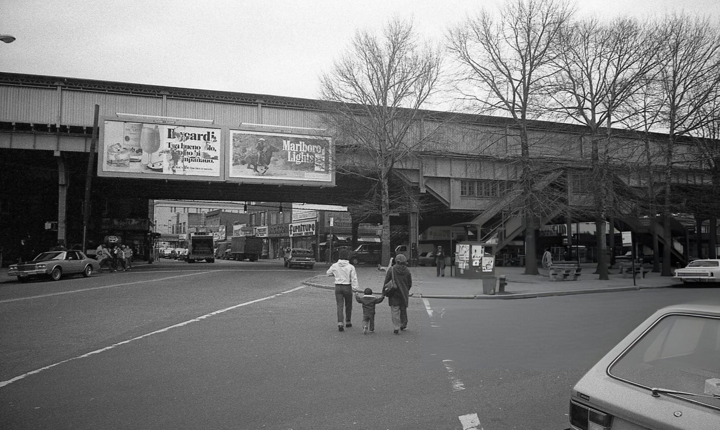 Two People Walk A Child Across The Intersection Of National Street And Roosevelt Avenue Near An Elevated Subway Line In Corona, Queens, 1982.