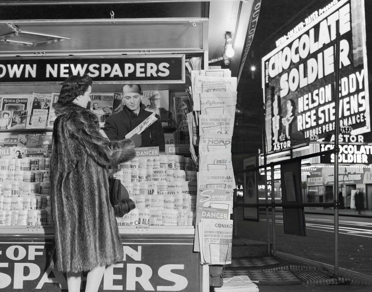 A Woman Wearing A Fur Coat Purchases A Newspaper From A Vendor At A Newsstand In Times Square, 1941.