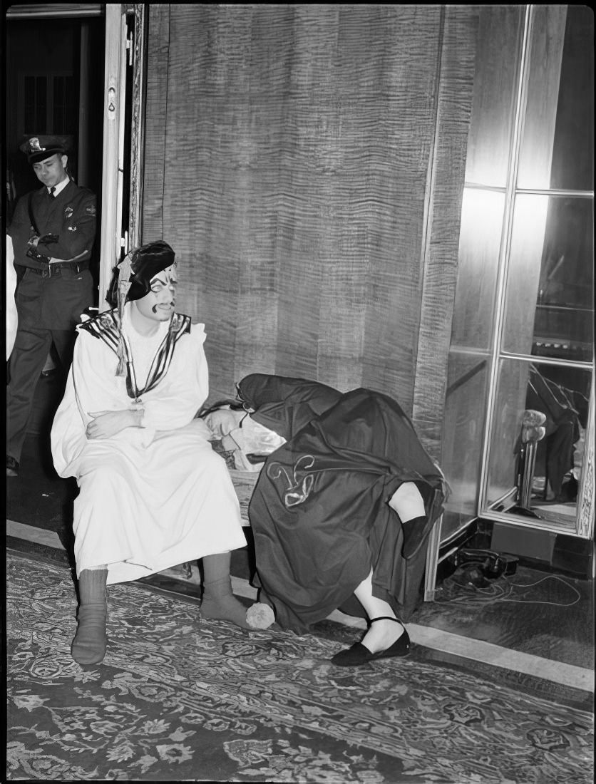 A woman sleeping on a small couch as a man in costume sits by her, 1951.