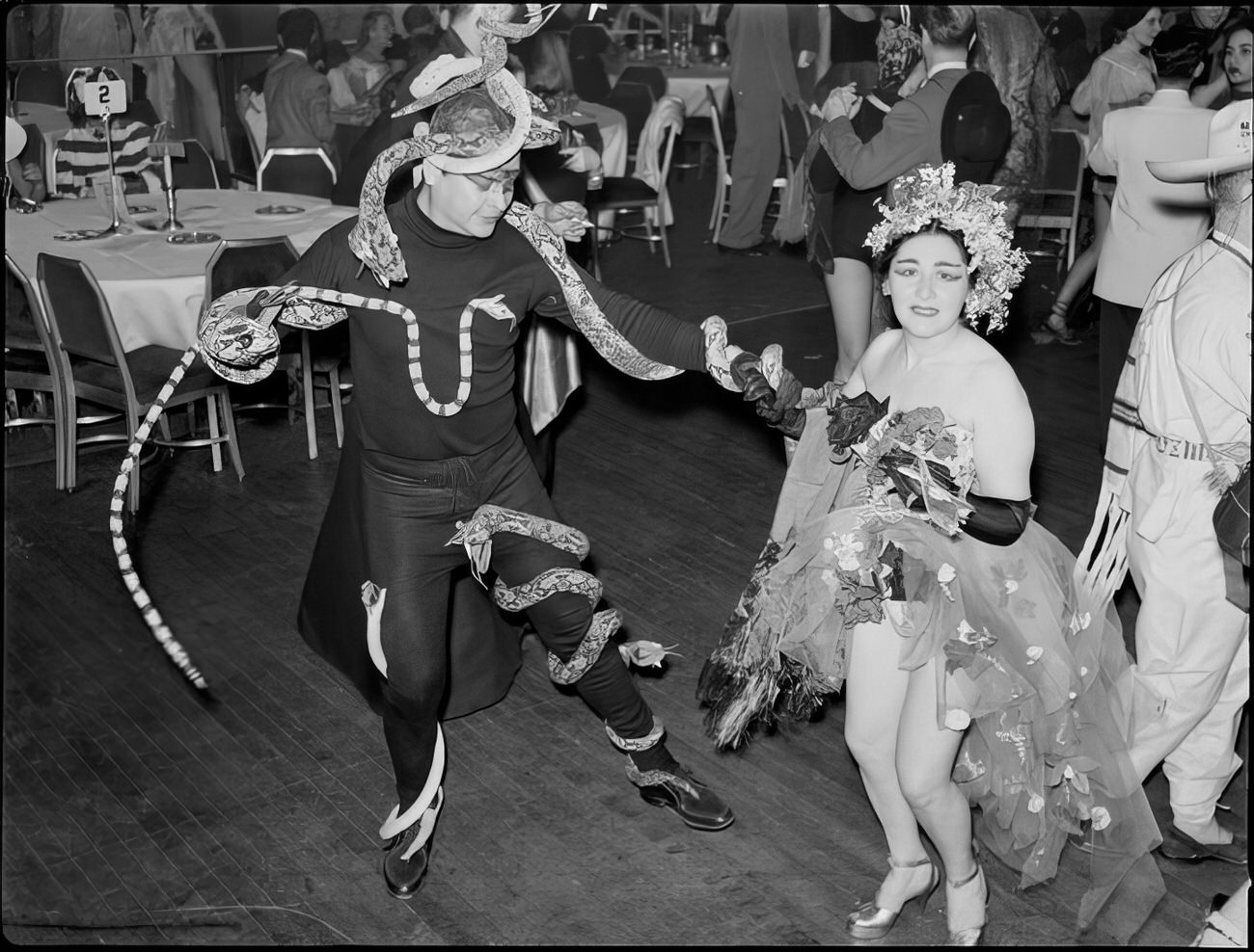 A man with fake snakes attached to him dancing with a woman wearing a headpiece at the ball, 1951.