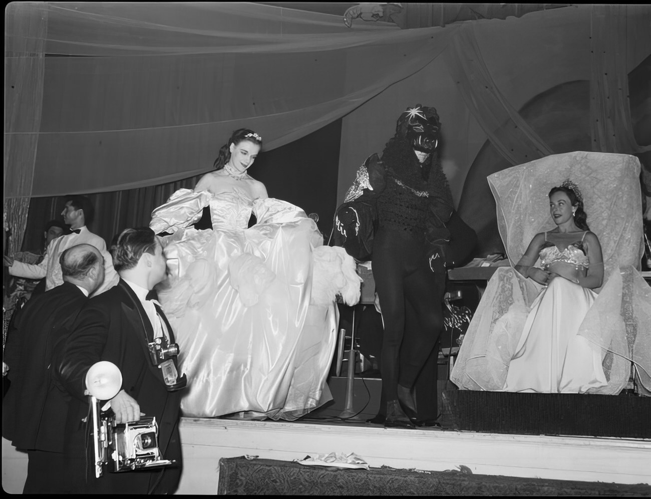 Two costumed ball guests walking across the stage as a woman watches, 1951.