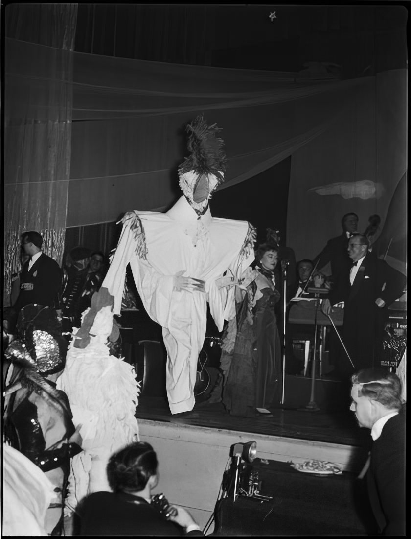 A woman leading a person in an elaborate costume across the stage, 1951.