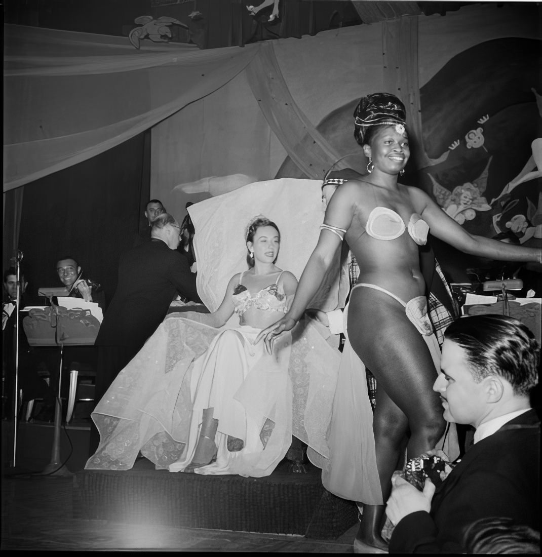 A costumed woman walking across the stage as another woman watches, 1951.