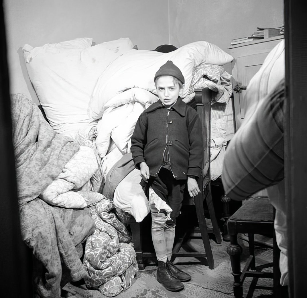 Child standing in front of piled-up furniture and bedding, 1936.