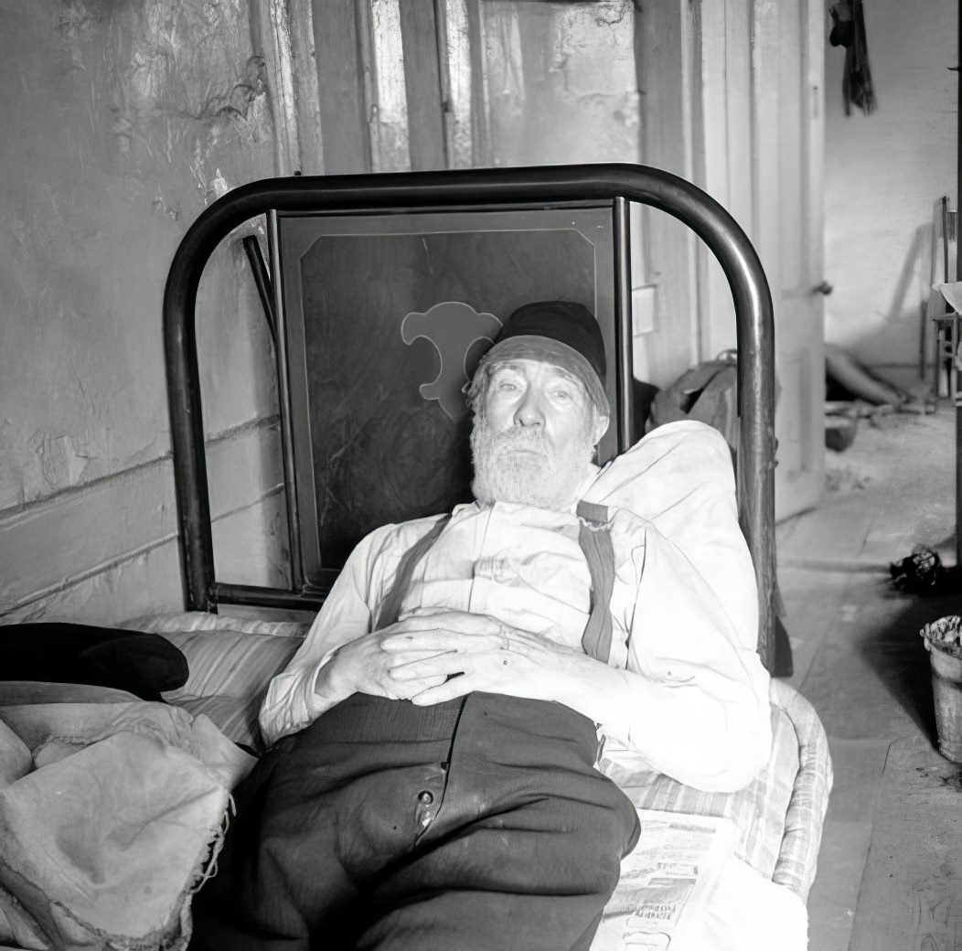 Bearded man resting on unmade bed, 1935.