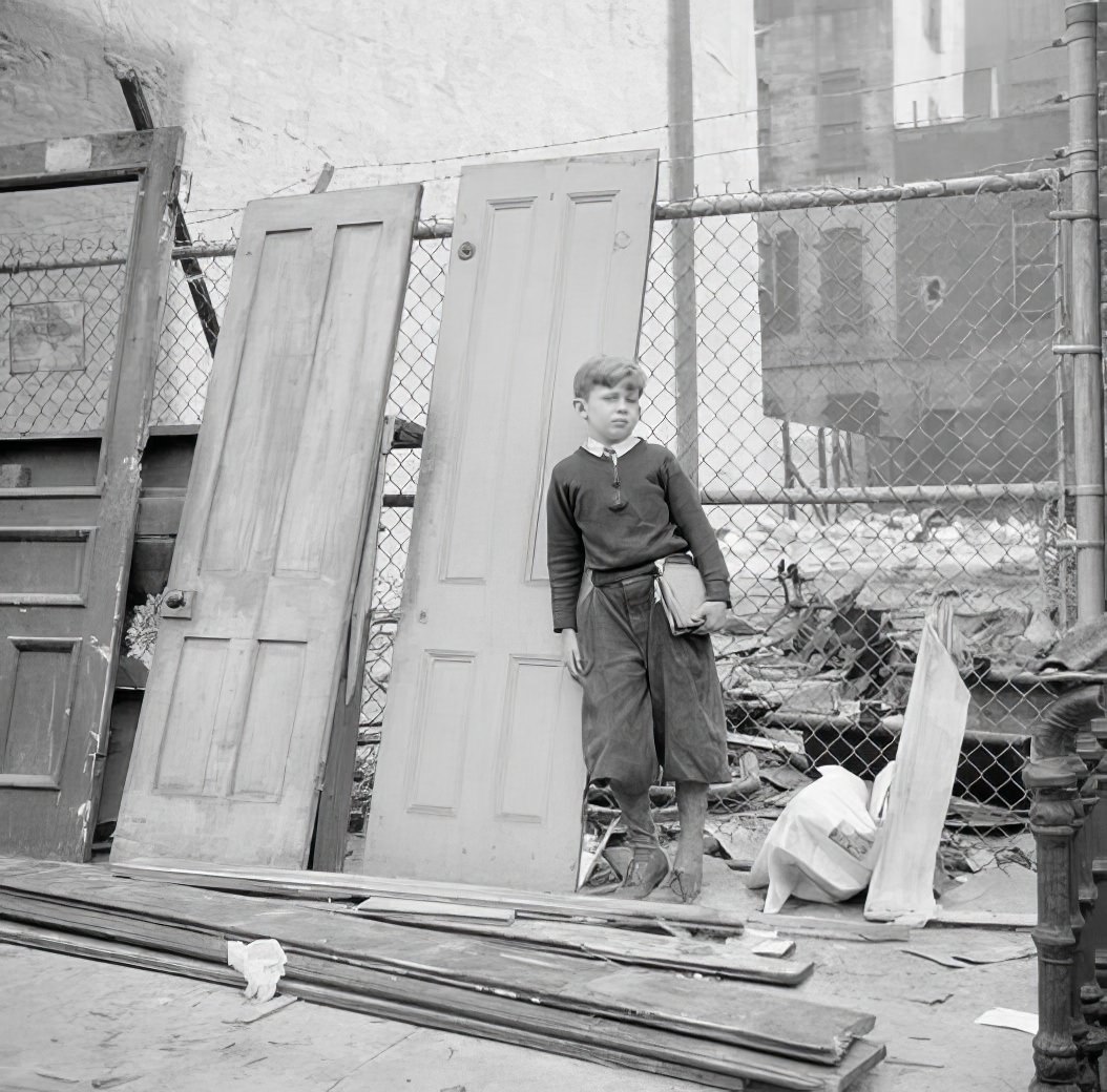 Schoolboy and old doors against wire fence, 1935.