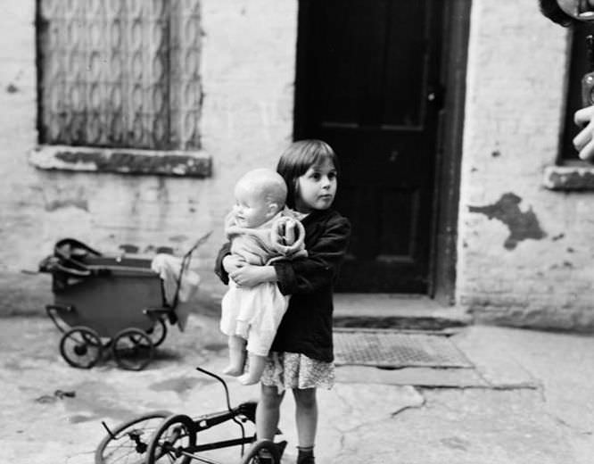 Little girl with doll and tricycle in front of shabby exterior, 1936.