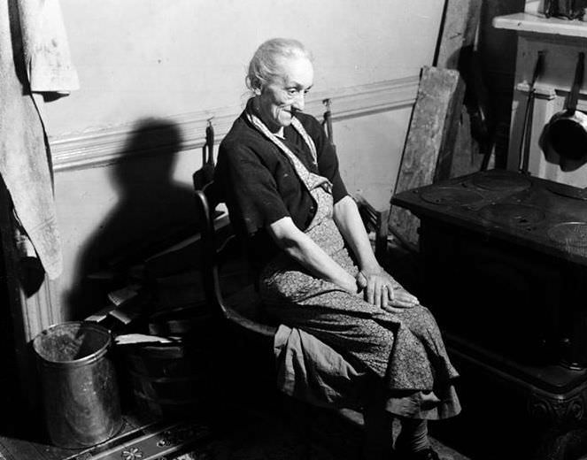 Old lady seated beside old-fashioned stove, 1936.