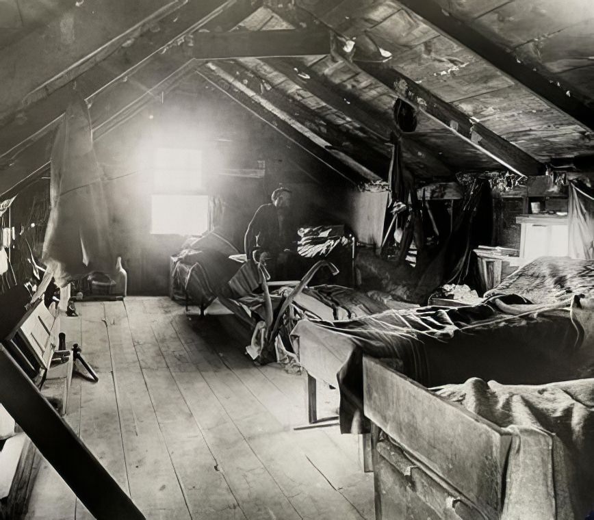 Man seated on bed in attic living quarters, circa 1895.