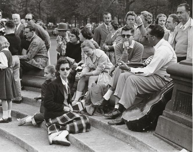 A woman in a plaid skirt and sunglasses looking at the photographer on the fountain steps, Washington Square Park, 1956.