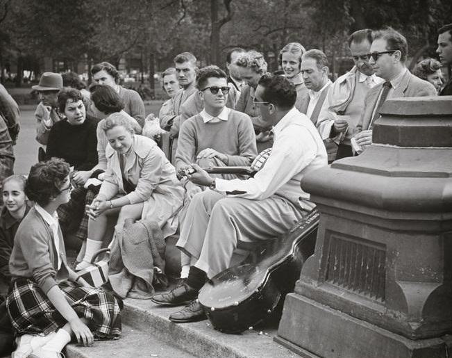 A woman in a plaid skirt watching a banjo player on the fountain steps, Washington Square Park, 1956.