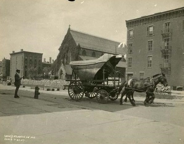 Subway construction site at the intersection of Atlantic Avenue and 4th Avenue. Image includes a horse-drawn wagon in right foreground, one man standing and a fire hydrant in the left foreground, and an unidentified church in the background.