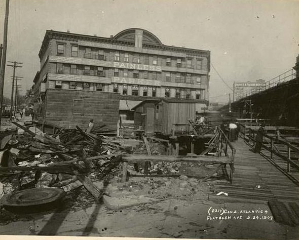 View of subway construction site at the intersection of Atlantic Avenue and Flatbush Avenue. Image includes a large, commercial or office building, located at 124 Flatbush Avenue, with a large sign, 'Painless Parker,' in the backgound. 1907.