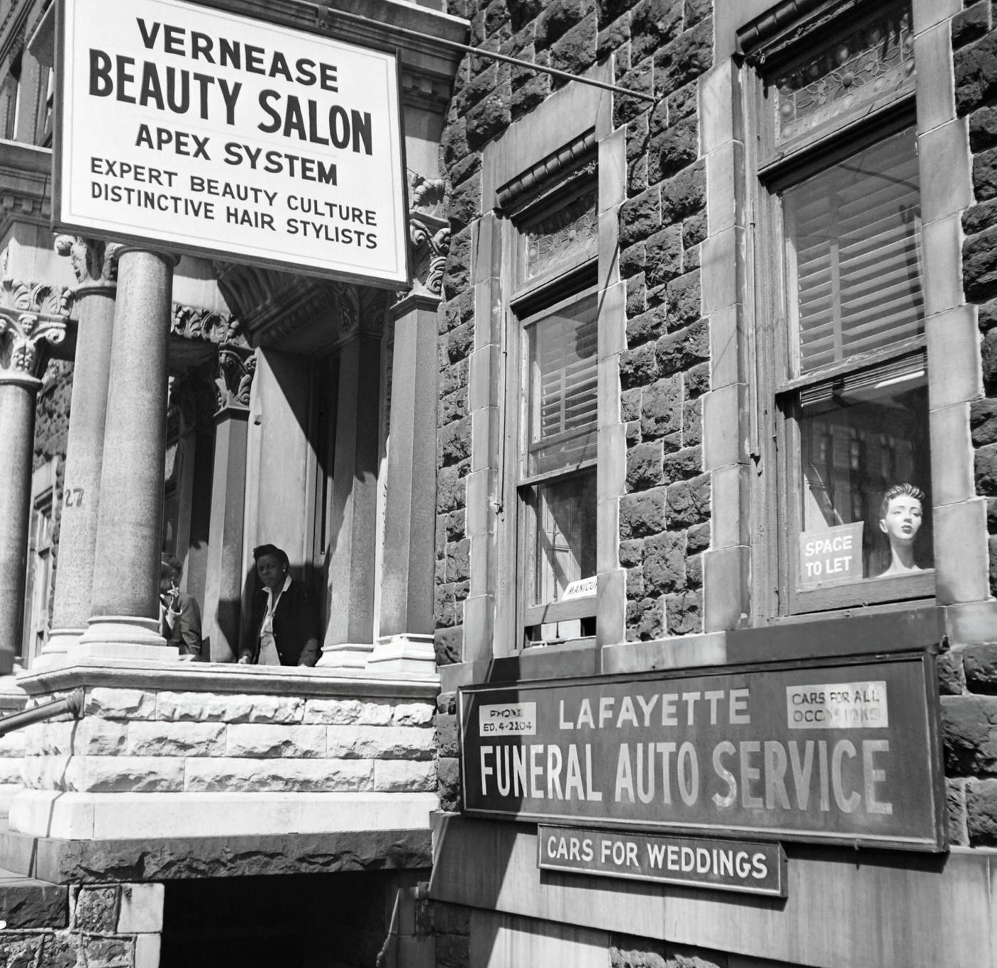 Two women stand in front of a beauty salon and a funeral and wedding cars service in Harlem, 1946.