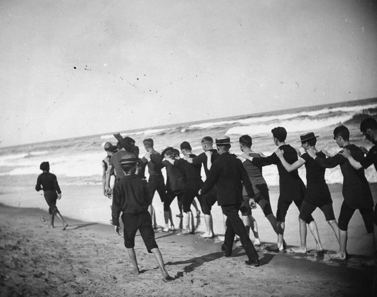 A group of young male bathers walk single file along the beach, 1886.
