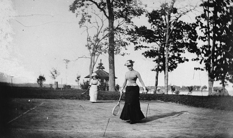 Two well-dressed women playing lawn tennis next to a lake, New York, 1880s.