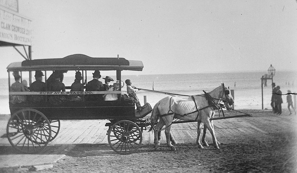 A young black boy pulling the reins of a horse drawn coach owned by the Surf Ave. Stage Co., alongside the boardwalk at Coney Island, 1884.