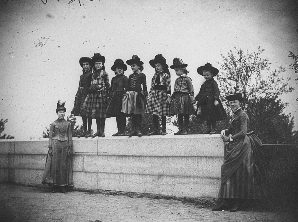Seven young girls including Isabel and Nellie Harter, Nellie and Edie Dwight, Zelma Levison and Edie Swan standing on a wall with two women, New York, 1886.