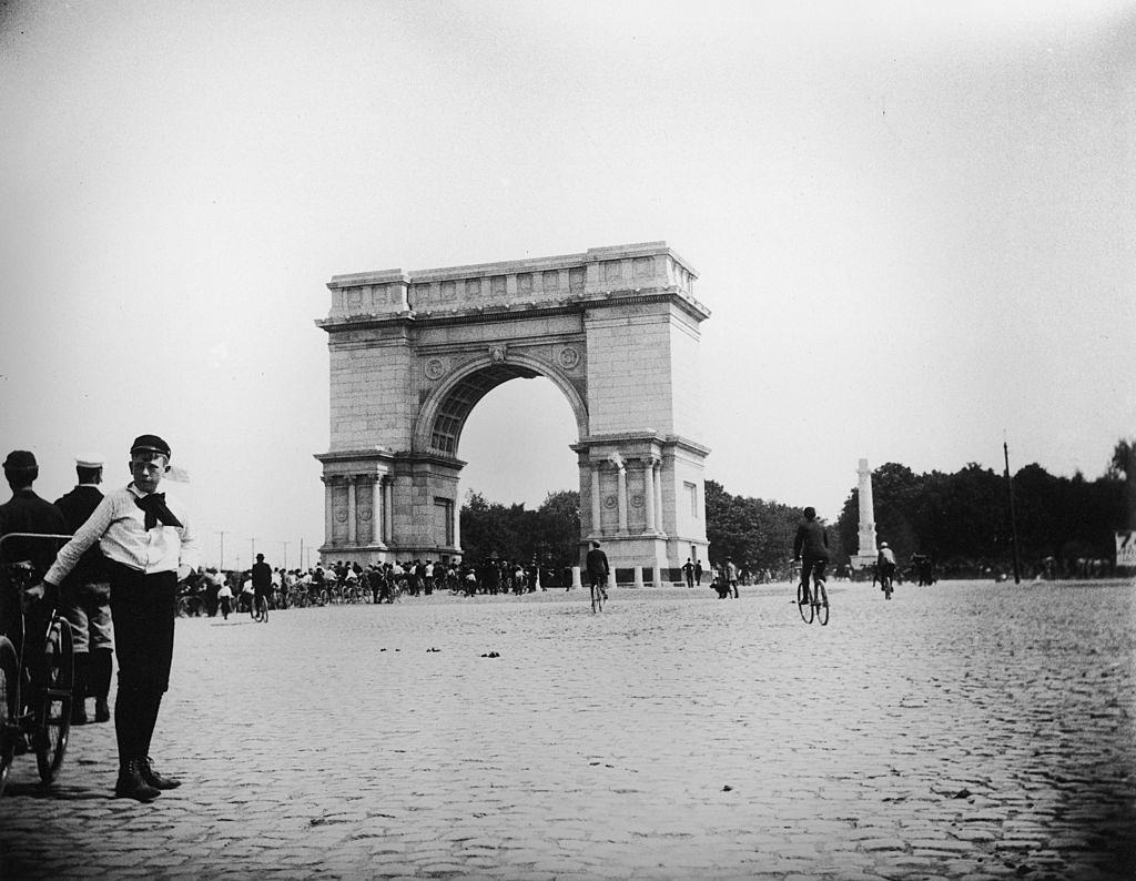 A boy leans against a bicycle in the foreground as people in the background ride their bikes through the arch at Prospect Park during a bicycle parade, Brooklyn, New York, 1895.