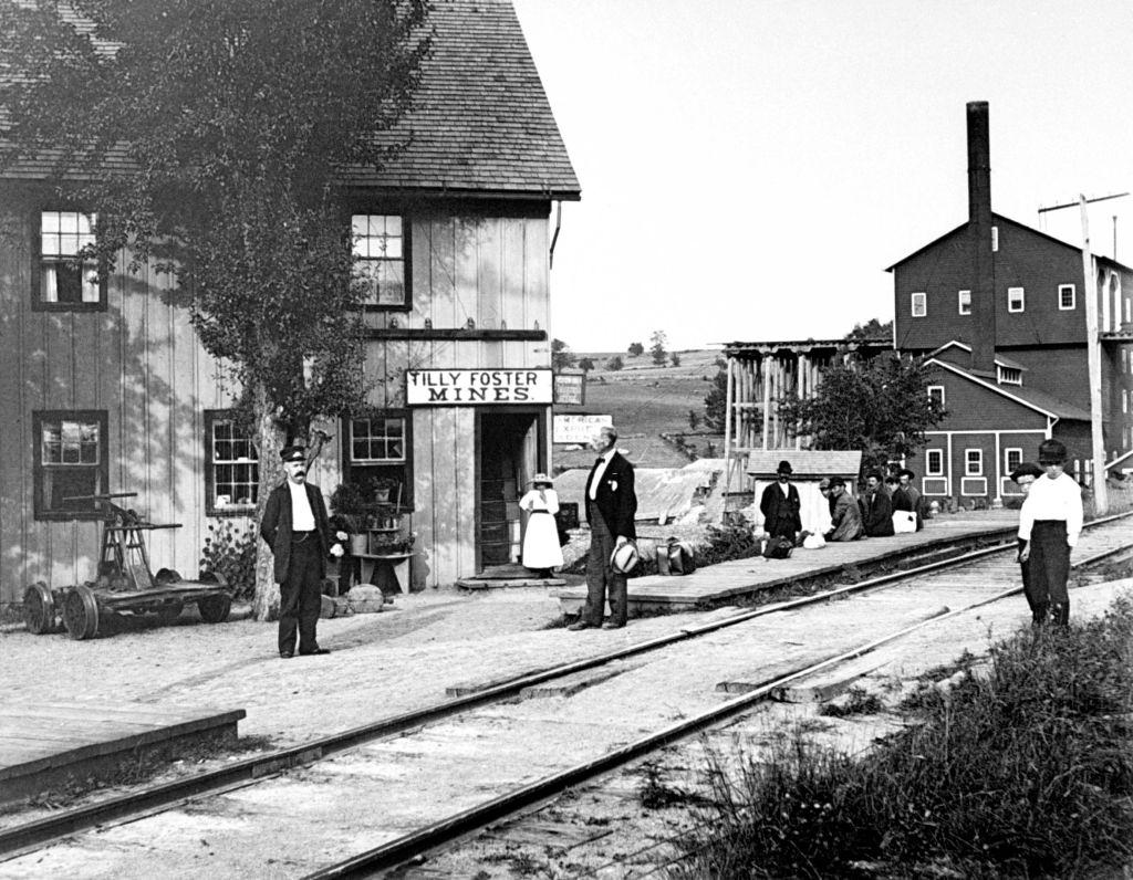 People hanging around outside railroad station, which has signs on it saying Tilly Foster Mines, New York, 1889.