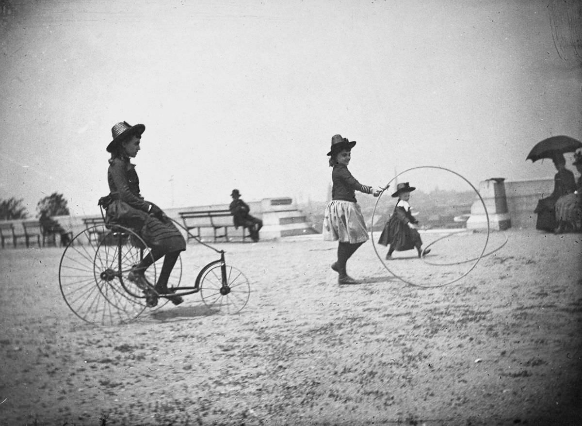 Isabel Harter rides a tricycle while her sister Nellie rolls a hoop in Fort Greene, 1886.