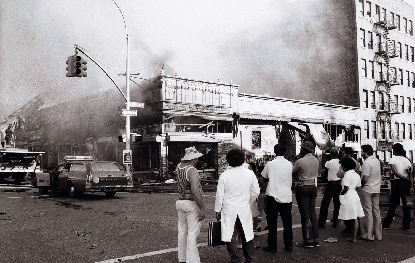 People look on as firemen put an end to arsonist's handiwork on Marmion Ave. in Brooklyn during the blackout, 1977.