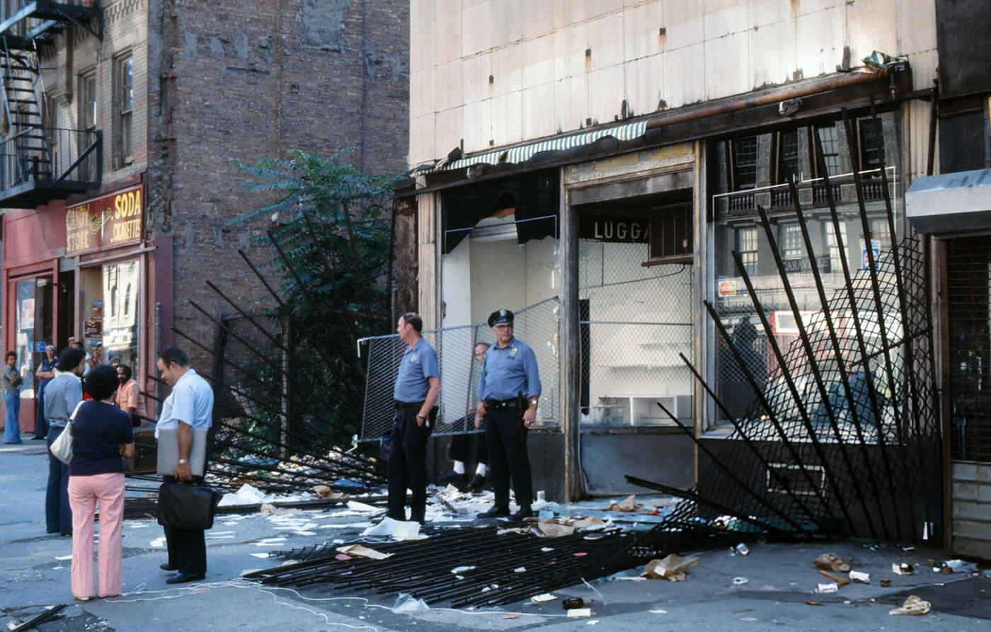 Police officers and passersby stand in front of a damaged store front, looted during the New York City blackout, Brooklyn, 1977.