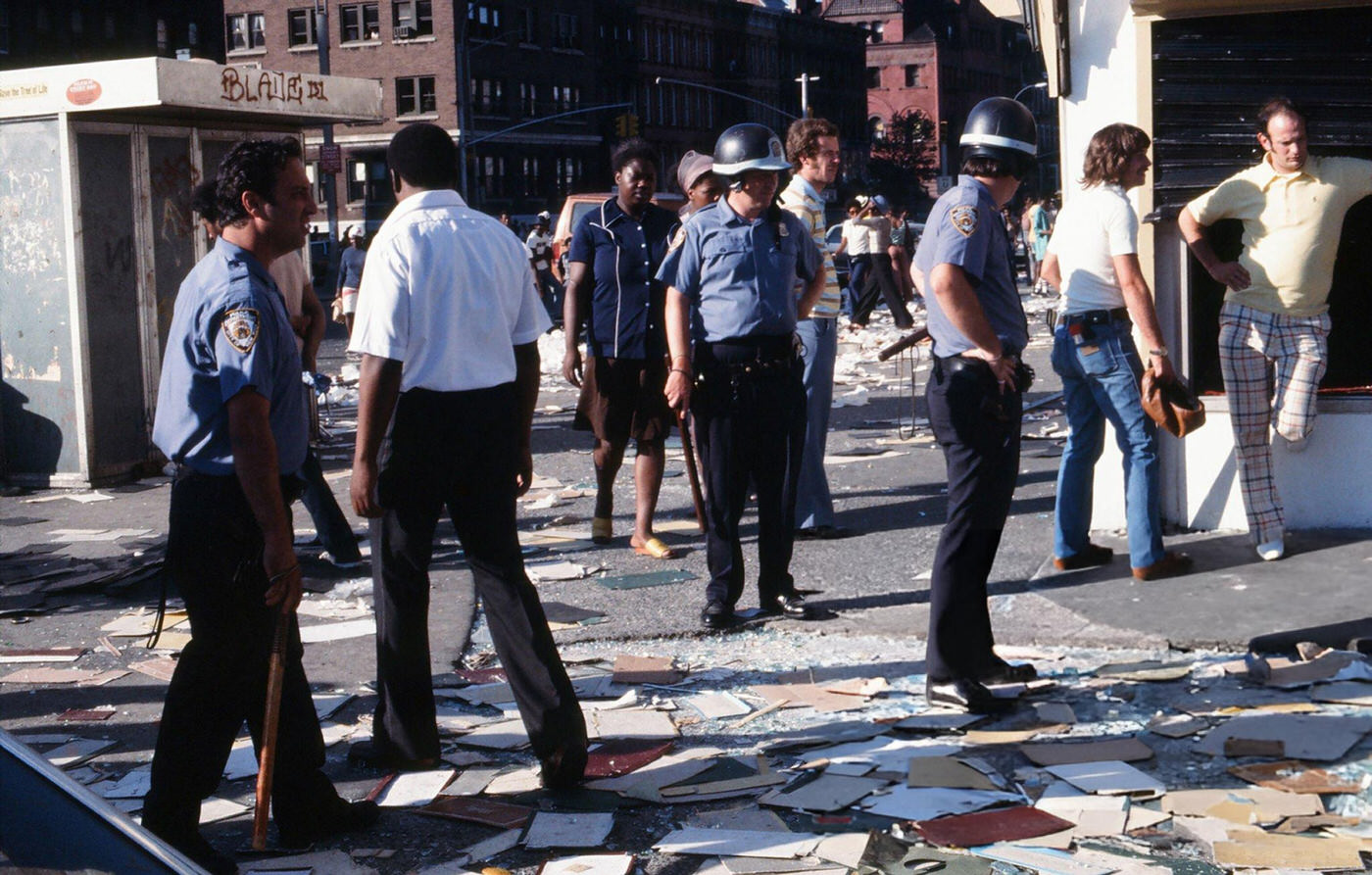 Police officers and passersby stand in front of a damaged store front, looted during the New York City blackout, Brooklyn, 1977.