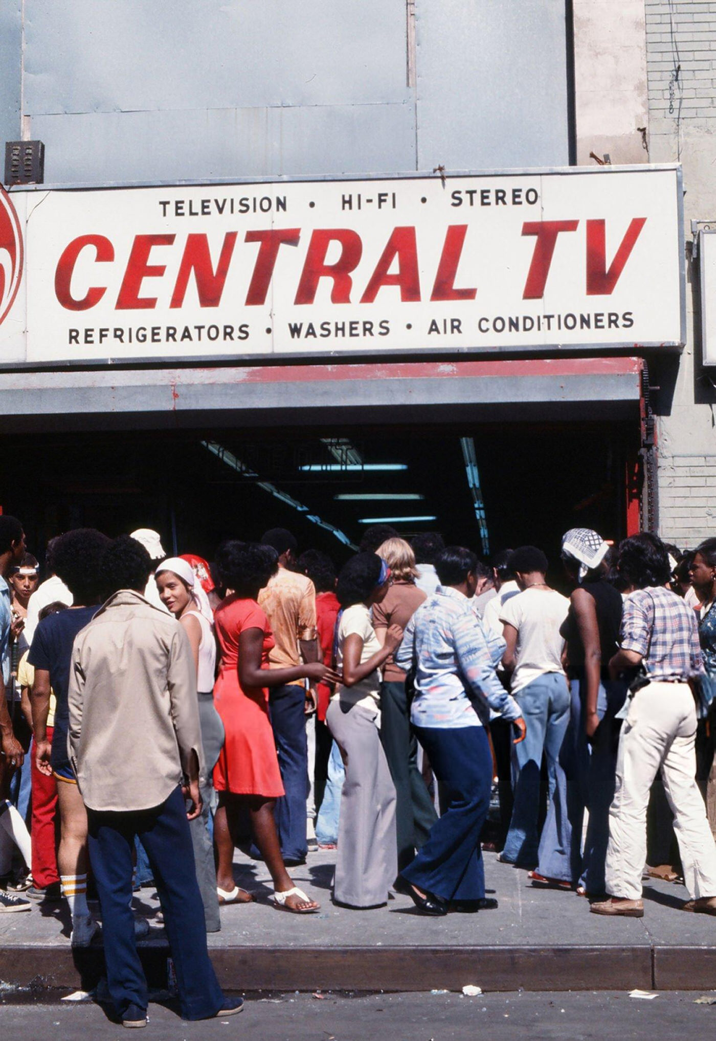 Passersby look into a damaged appliance store, looted during the New York City blackout, Brooklyn, 1977.