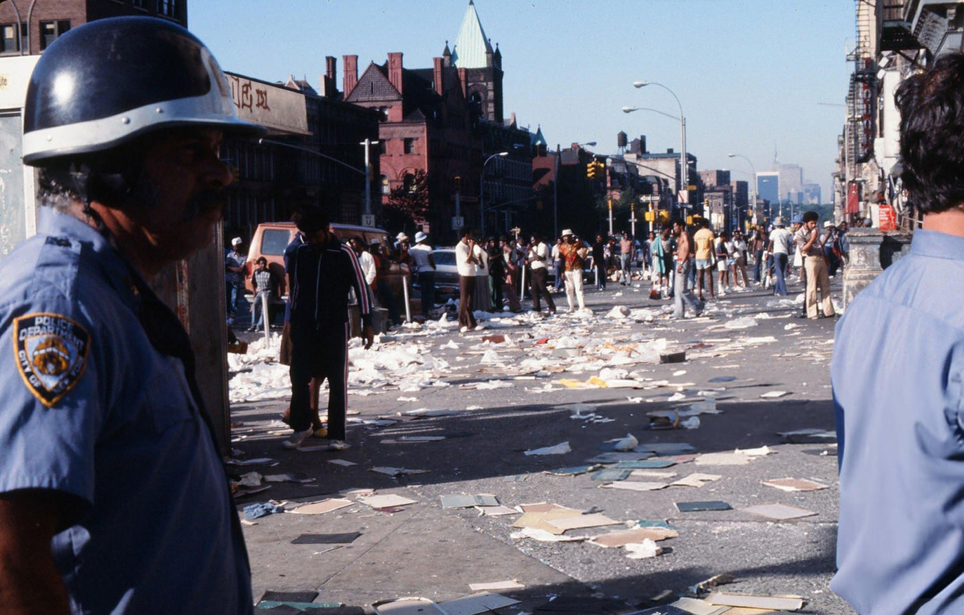 Police officers and passersby stand in front of a damaged store front, looted during the New York City blackout, Brooklyn, 1977.