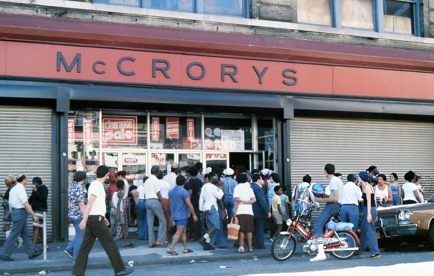 Passersby stand in front of a damaged store front, looted during the New York City blackout, Brooklyn, 1977.