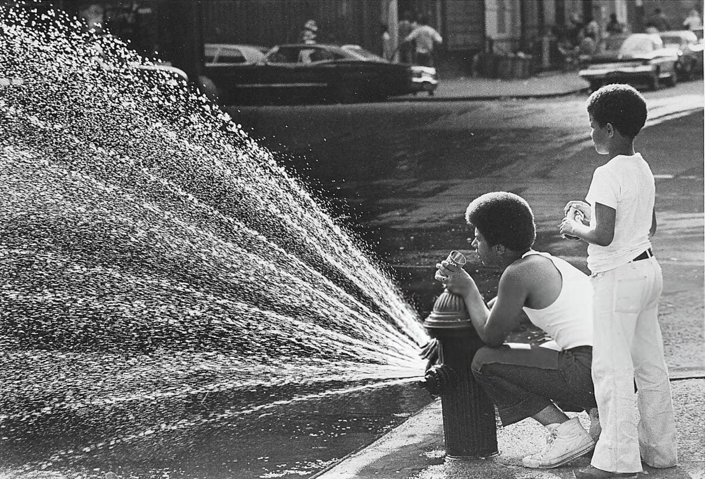 A teenager and a younger child stand near the sprinkler of an open fire hydrant in Harlem in the aftermath of the blackout, 1977.