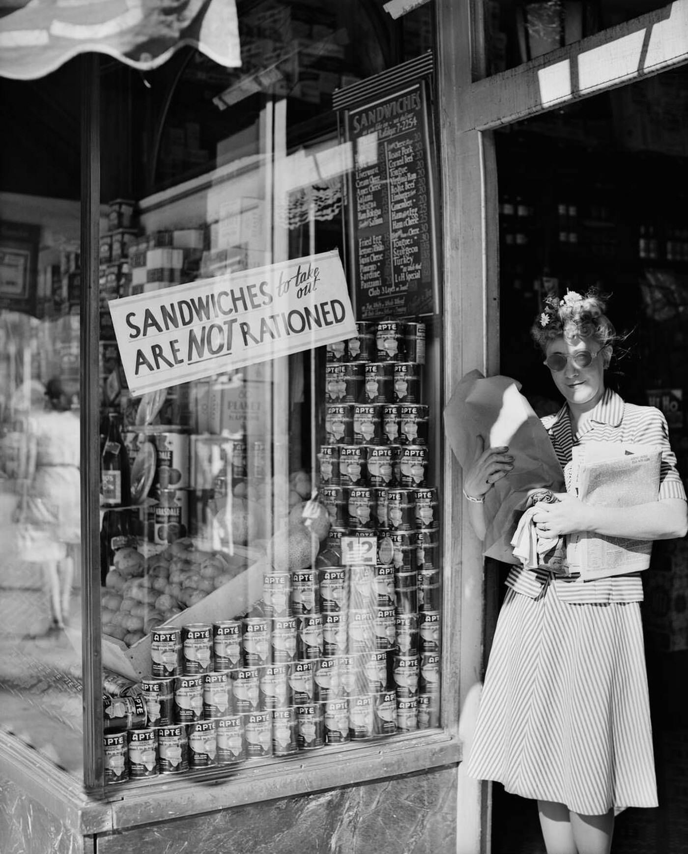 Woman carrying packages in door of New York City delicatessen displaying a sign "Sandwiches to Take Out Are Not Rationed", 1940s.