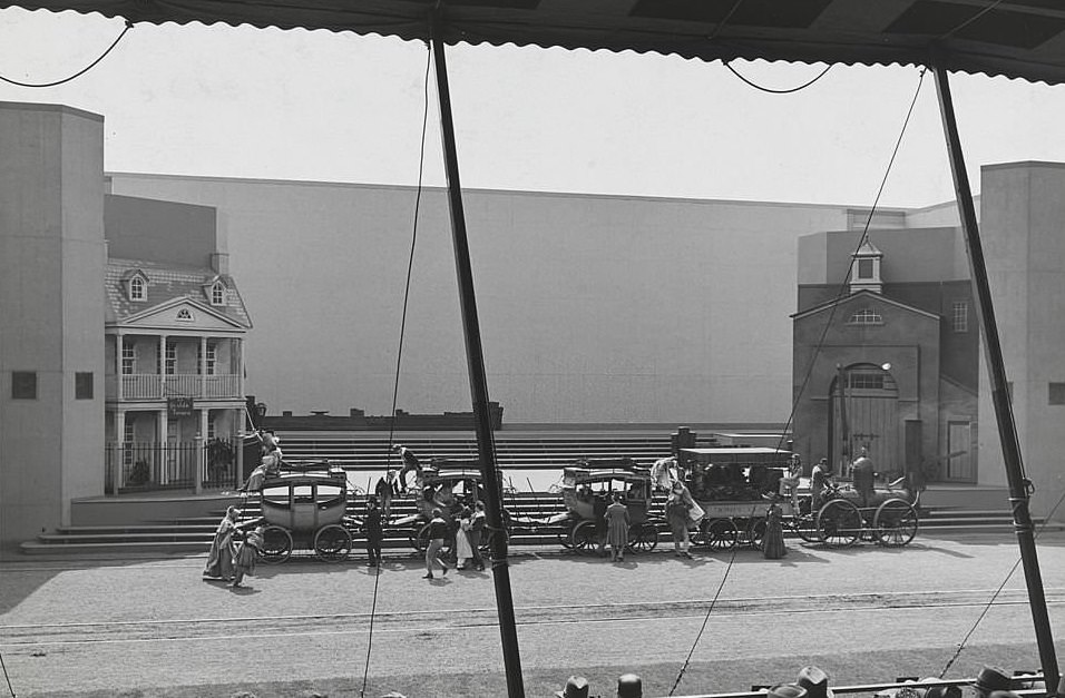 An outdoor performance featuring actors in costume on a set featuring a small steam engine pulling a passenger car and stagecoaches.