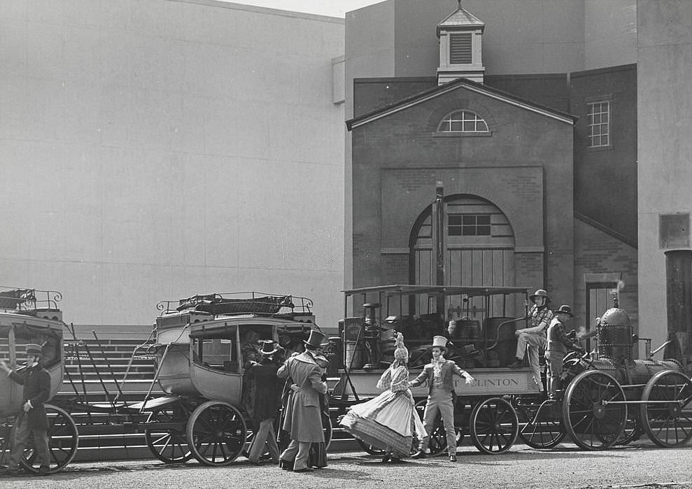 An outdoor performance featuring actors in costume on a set featuring a small steam engine pulling a passenger car and stagecoaches.