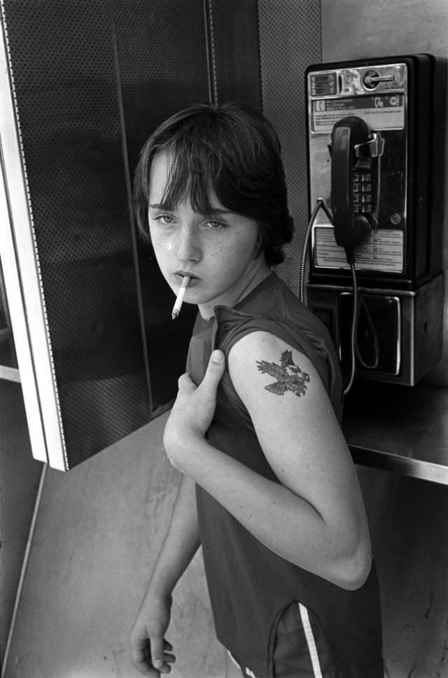 A young hustler smokes in a telephone booth, surrounded by signs of urban decay.