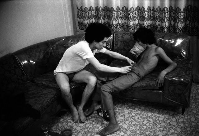 A 16-year-old sniffs glue from a paper bag while an older hustler undresses him in 1979—illustrating the mix of addiction, abuse, and exploitation.
