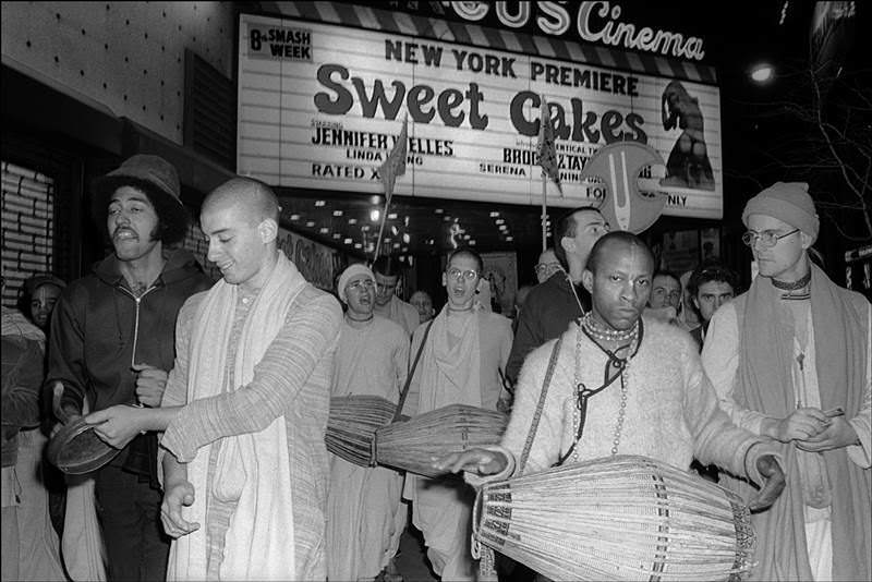 Hare Krishna followers chant and play instruments under the marquee of an adult theater showing Sweet Cakes, Times Square, 1976.